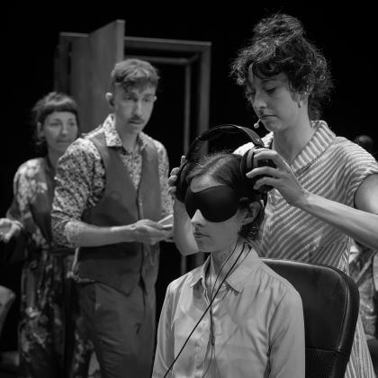 A woman is putting headphones on another woman who wearing blindfolds. Two people, on their right side, are looking at them.